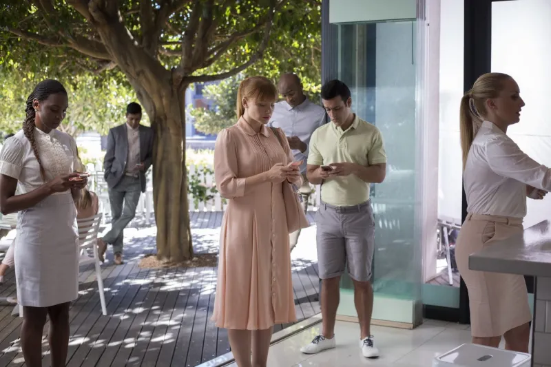 Женщина в розовом платье с тревогой смотрит в свой смартфон на залитой солнцем террасе, в то время как другие люди вокруг нее также поглощены своими телефонами, иллюстрируя социальную изоляцию.