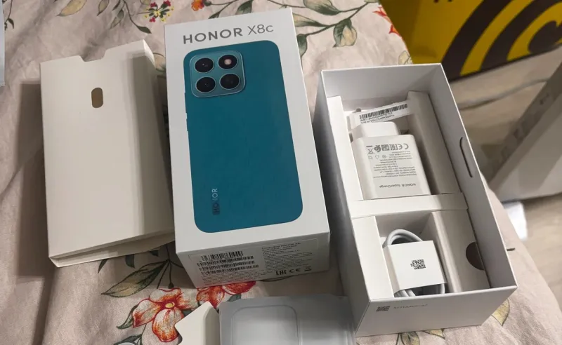 Распаковка смартфона HONOR X8c: на фото показана коробка, бирюзовый телефон и комплектные аксессуары — зарядное устройство SuperCharge и кабель.