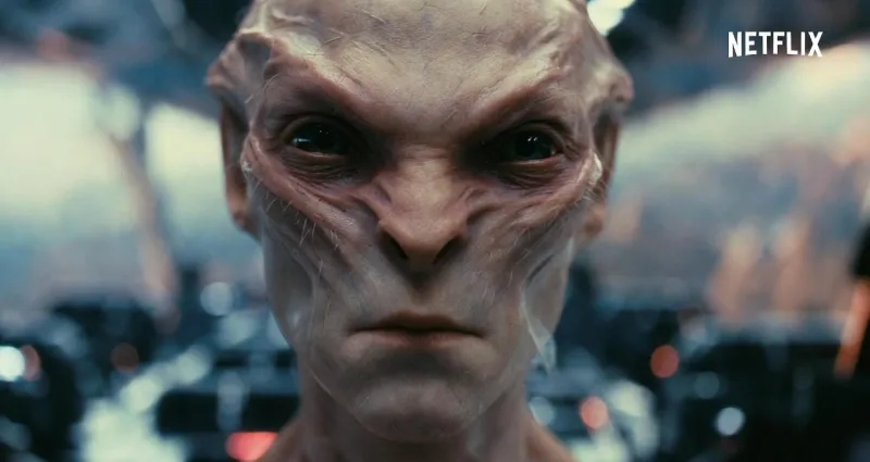 Крупный план лица бледного, безволосого гуманоидного пришельца с серьезным выражением лица, глубоко посаженными глазами и выраженными скулами. В правом верхнем углу виден логотип Netflix. 10 лучших научно-фантастических сериалов Netflix, которые стоит посмотреть