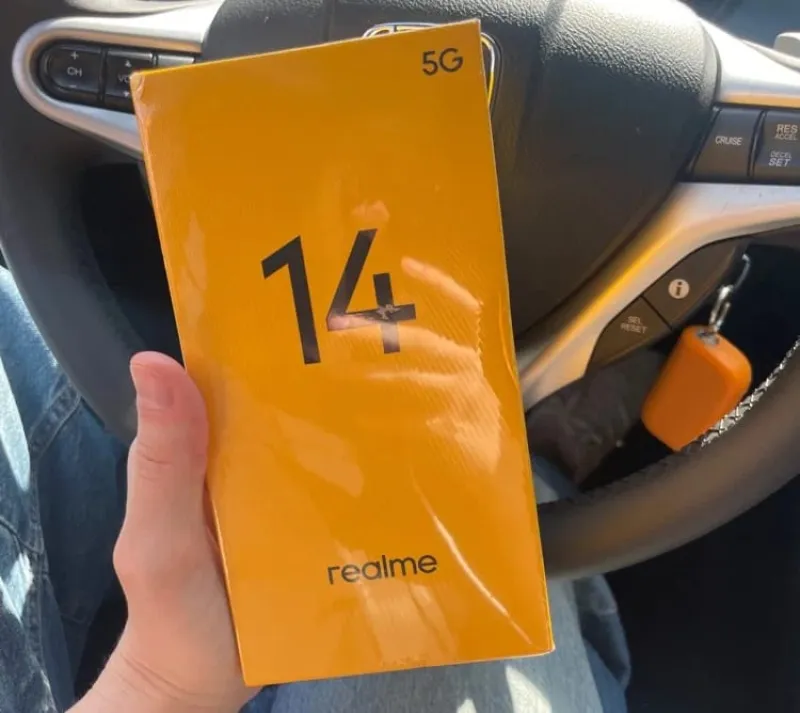 Рука держит новую, запечатанную в пленку, желтую коробку смартфона Realme 14 5G в салоне автомобиля.