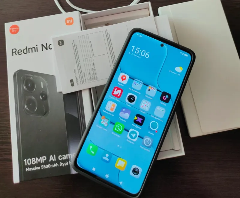 Смартфон Xiaomi Redmi Note 14 с включенным экраном, на котором видны приложения, лежит на фоне своей коробки с указанием характеристик «108MP AI camera» и «Massive 5500mAh».