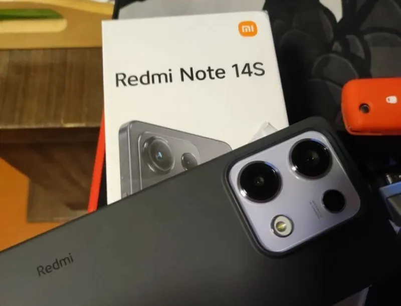 Смартфон Redmi Note 14S в темном корпусе с крупным модулем камеры, лежащий на фоне своей белой коробки.
